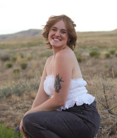 Sarah, a website copywriter, posing in an open field and smiling. She is wearing a white strapless top and black jeans, and has a bird tattoo on her upper arm.